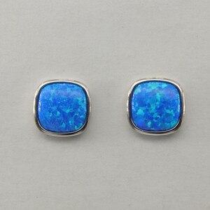 Puede incluir: Un par de pendientes cuadrados con ópalos azules. Los ópalos presentan una mezcla de tonos azules claros y oscuros, engastados en un marco de metal plateado. Los pendientes se muestran sobre un fondo gris claro.