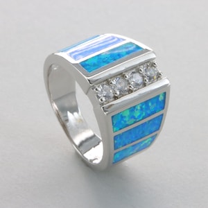 May include: A silver ring with a rectangular face featuring three horizontal bands of blue opal and three small clear stones. The ring has a polished finish and a modern design.