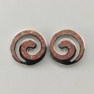 Puede incluir: Un par de pendientes de botón plateados. Cada pendiente presenta un diseño circular con una incrustación en espiral de material iridiscente, rojo-anaranjado. Los pendientes están sobre un fondo liso y claro.