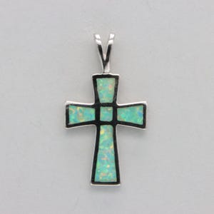 Puede incluir: Un colgante de cruz de plata con una incrustación de ópalo azul claro. La cruz está hecha de plata de ley y tiene un acabado pulido.