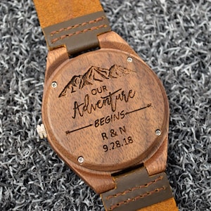 May include: Wooden watch with a brown leather strap. The watch face is engraved with a mountain scene and the words "OUR Adventure BEGINS" along with the initials "R & N" and the date "9.28.18".
