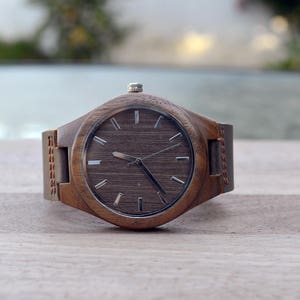 May include: A wooden wristwatch with a brown face and a matching leather band. The watch has silver-colored hands and markers. The watch is displayed on a light-colored wooden surface.