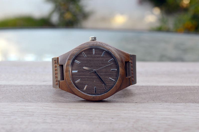 May include: A wooden wristwatch with a brown face and a matching leather band. The watch has silver-colored hands and markers. The watch is displayed on a light-colored wooden surface.