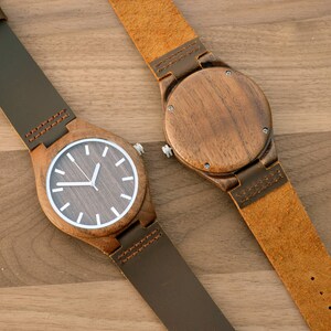 May include: Two wooden wristwatches with brown leather straps. One watch face is visible, featuring a dark wood grain dial with white markers and hands. The other watch shows the back of the wooden watch case and a tan leather strap.