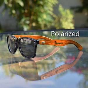 May include: Polarized sunglasses with a black frame and wooden arms. The arms are engraved with the name "Thompson". The sunglasses are reflected in the surface below. The text "Polarized" is displayed above the sunglasses.