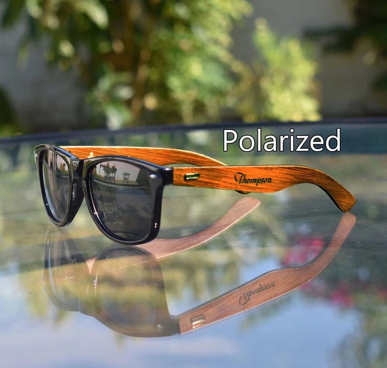 May include: Polarized sunglasses with a black frame and wooden arms. The arms are engraved with the name "Thompson". The sunglasses are reflected in the surface below. The text "Polarized" is displayed above the sunglasses.