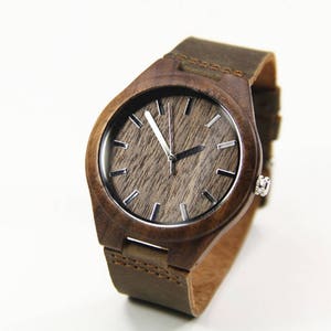 May include: A wooden wristwatch with a brown leather band. The watch face has a wood grain pattern with silver-colored hour markers and hands. The watch case is made of dark brown wood.