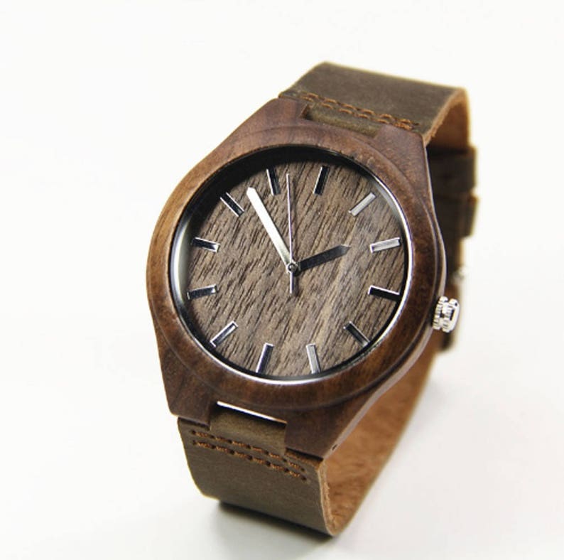 May include: A wooden wristwatch with a brown leather band. The watch face has a wood grain pattern with silver-colored hour markers and hands. The watch case is made of dark brown wood.