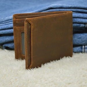 May include: A brown leather wallet with a stitched edge and a card slot. The wallet is sitting on a white fuzzy surface.