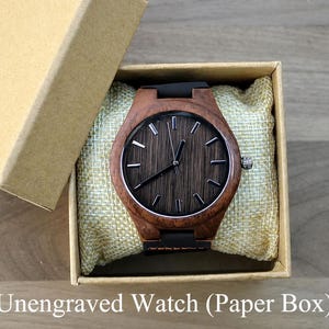 May include: A wooden watch with a dark brown face and a black band rests in a beige box with a matching lid. The watch has silver-colored hands and markers. The watch is presented on a beige cushion.