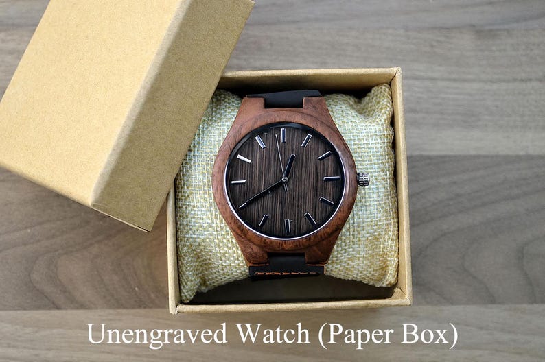 May include: A wooden watch with a dark brown face and a black band rests in a beige box with a matching lid. The watch has silver-colored hands and markers. The watch is presented on a beige cushion.