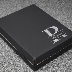 May include: A black box with a white stitched border. The box has a silver monogram with the letter "P" and the name "Reginald" engraved on the top. The box is sitting on a gray surface.