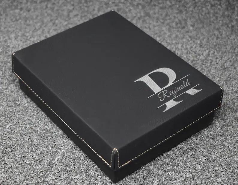 May include: A black box with a white stitched border. The box has a silver monogram with the letter "P" and the name "Reginald" engraved on the top. The box is sitting on a gray surface.
