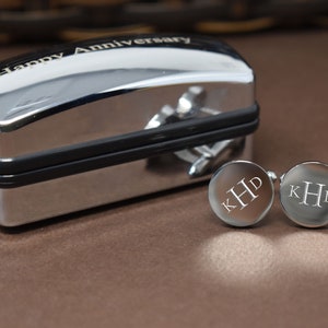 May include: A silver cufflinks box with the inscription "Happy Anniversary" on the top. The box is open and two silver cufflinks with the initials "KHD" engraved on them are visible.