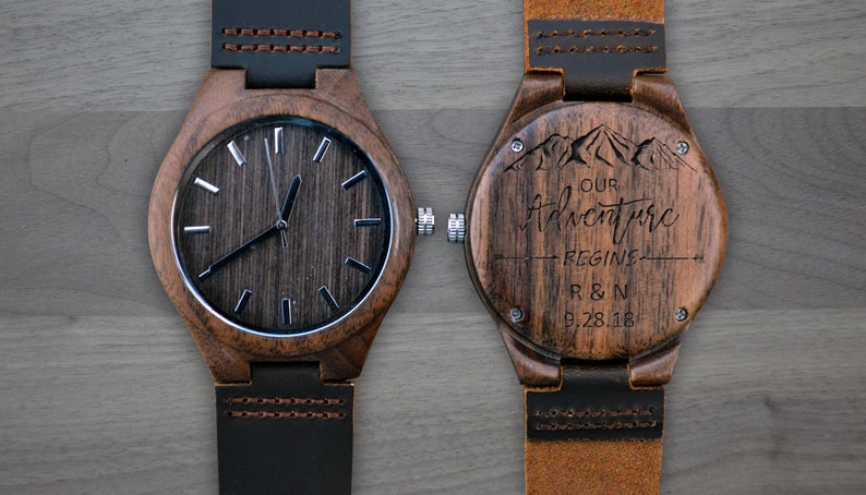 May include: Wooden wristwatch with a dark wood grain face and a black leather strap. The watch face has silver-colored hour markers. The back of the watch is engraved with the words "Our Adventure Begins", mountains, and the initials "R & N" with the date "9.28.18".