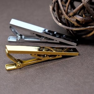 May include: Two metal tie clips, one silver and one gold, are displayed on a brown surface. The gold tie clip has the words "World's Best Dad" engraved on it. A decorative woven ball is in the background.
