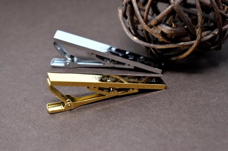 May include: Two metal tie clips, one silver and one gold, are displayed on a brown surface. The gold tie clip has the words "World's Best Dad" engraved on it. A decorative woven ball is in the background.