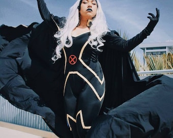 May include: A woman dressed as Storm from the X-Men comics. She is wearing a black and gold costume with a long black cape. Her hair is white and styled in a long, flowing wig. She is posing with her arms outstretched, as if she is about to unleash a powerful storm.