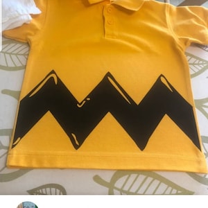 K&ouml;nnte beinhalten: Gelbes Poloshirt mit einem schwarzen Zickzackmuster am unteren Rand. Das Shirt hat einen Kragen und Kn&ouml;pfe.