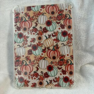 Op de afbeelding: Transparante tablet hoes met een herfstthema patroon. Het ontwerp bevat pompoenen in oranje, wit en groen, samen met rode rozen, zonnebloemen en herfstbladeren. De hoes heeft een transparante beschermende rand.