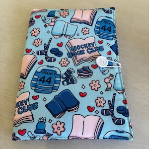 May include: A light blue book cover featuring a pattern of hockey jerseys, books, hockey sticks, and flowers. The cover has the text "HOCKEY BOOK CLUB" and a white button closure.