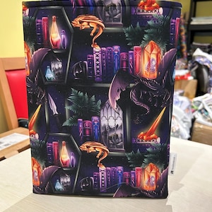 May include: A black book cover with a repeating pattern of dragons, books, crystals, and potions. The background is a dark blue with stars. The text "Bookish" is printed on the bottom right corner.
