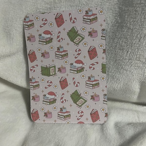 以下が含まれることがあります： ピンクの背景に、本、キャンドル、キャンディーケーン、白い花が繰り返しパターンで描かれています。本は積み重ねられており、緑色の表紙に「Tis the Season」という文字が書かれています。キャンドルはピンクと白です。キャンディーケーンは赤と白です。花は白で、中心は黄色です。