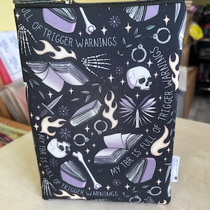 Puede incluir: Bolsa negra con cremallera con un patrón repetido de libros, cráneos, huesos, esposas, llamas y estrellas. El texto "My TBR is full of trigger warnings" se repite en la bolsa.