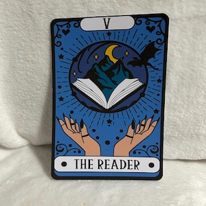 May include: A blue tarot card with a black border and a white circle in the centre. The circle contains the text "THE READER" in black. The card also features a crescent moon, a mountain, a book, and two hands with black fingernails. The top of the card has a white rectangle with the number "V" in black.