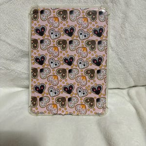 Puede incluir: Una funda rectangular para teléfono con borde transparente. La funda presenta un patrón repetido de corazones, huellas de patas y estrellas en tonos de negro, marrón y blanco sobre un fondo rosa.