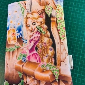 Op de afbeelding: Een witte stoffen boekhoes met een kleurrijke illustratie van Rapunzel uit de Disney-film Tangled. Rapunzel staat in een torenraam met haar lange blonde haar en een groene kameleon op haar schouder. De tekst "bookthetique" is op de hoes gedrukt.