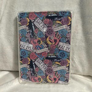 May include: Clear protective case with a colorful design featuring roses, license plates with "KAZ 2Y5", a black car, and pentagrams. The case has a repeating pattern with the words "The Family Business".