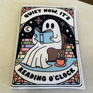 May include: A book cover with a whimsical design featuring a cartoon ghost reading a book while seated in a chair. The cover has the text "QUIET NOW, IT'S READING O'CLOCK" and a button closure.