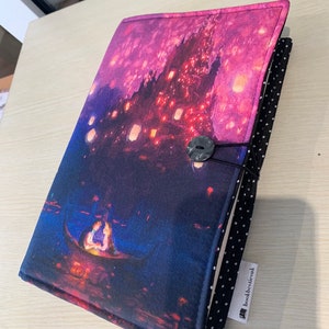 Può includere: Una copertina di libro viola e blu con una scena di un castello illuminato da lanterne e una coppia in barca. La copertina ha un tessuto a pois bianco e nero sul retro con un'etichetta bianca che dice "bookbestiesuk".