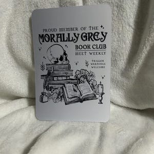 May include: A white card with black text and illustrations. The text reads "Proud member of the Morally Grey Book Club Meet Weekly Trigger Warnings Welcome". The illustration is of a skull, books, flowers, and a candle.