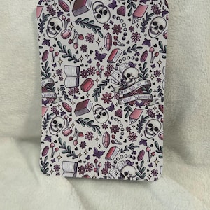 Puede incluir: Un objeto rectangular con un patrón repetido de calaveras, libros, cristales, flores y mariposas en blanco, morado y rosa. El texto "Trigger Warnings" y "You had me at" son visibles.