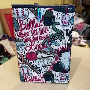 Puede incluir: Una bolsa rectangular con cremallera y un diseño colorido. El patrón incluye texto, pelotas de béisbol, cactus y una ilustración de vampiro. El texto incluye el nombre "Bella" y la frase "Where the Hell Have You Been Loca?" Un dije en forma de prisma cuelga de la cremallera.