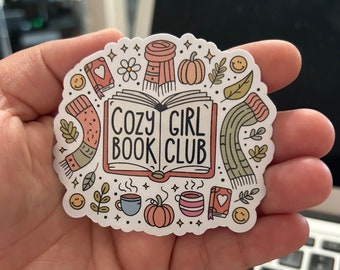 Op de afbeelding: Een witte sticker met een boek, een mok, een pompoen en een sjaal. De tekst op de sticker luidt "Cozy Book Club".