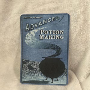 Könnte beinhalten: Ein blaues Buch mit dem Titel "Advanced Potion Making" von "Libatius Borage's" mit einer Illustration eines schwarzen Kessels.