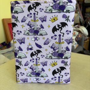 May include: A lavender book sleeve featuring a repeating pattern of black dragons, butterflies, swords, crowns, roses, and open books. The design includes purple and yellow accents. The sleeve is rectangular and appears to be made of fabric.