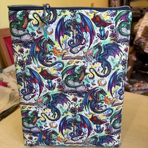 May include: A rectangular zippered bag with a fantasy dragon pattern. The bag features a repeating design of colorful dragons with open books, crystals, and floral elements on a white background. The zipper is dark blue.