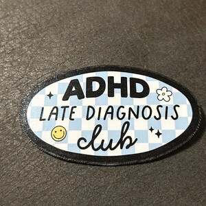 May include: A blue and white checkered oval patch with black text that reads "ADHD Late Diagnosis Club". The patch has a black border and features a smiley face, a flower, and two stars.