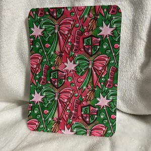 Könnte beinhalten: Ein grüner und rosa gemusterter Stoff mit einem sich wiederholenden Muster aus Schleifen, Zauberstäben, Besen und einem Wappen mit dem Text "Pink Goes Good With Green".