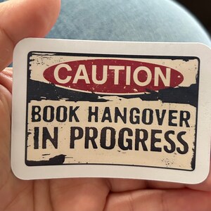 Puede incluir: Una pegatina blanca con un letrero de estilo vintage desgastado que dice "ATENCIÓN RESACA DE LIBRO EN CURSO". El letrero es rojo, negro y blanco.