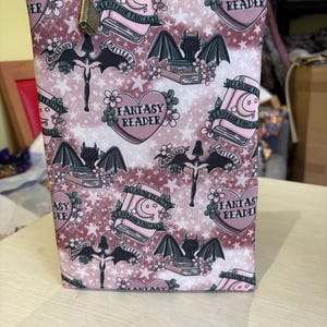 Può includere: Un astuccio rosa con cerniera con un motivo ripetuto di pipistrelli, libri, cuori e le parole "Fantasy Reader" e "Leaving Reality, Entering Fantasy." L'astuccio ha un tirazip dorato.