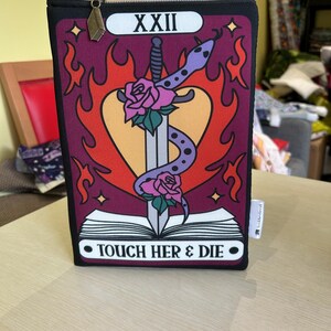 May include: Black zippered pouch with a tarot card design. The card is red, orange, and yellow with a heart, a sword, and a snake. The text on the card reads "XXII TOUCH HER & DIE".
