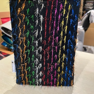 Op de afbeelding: Zwarte stoffen tas met een regenboog van kleurrijke metalen draden die aan de voorkant zijn genaaid. De tas heeft een label met de tekst "bookhoused" aan de zijkant.