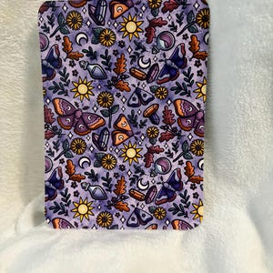 Könnte beinhalten: Rechteckige, lila Oberfläche mit einem sich wiederholenden Muster aus Motten, Sonnen, Monden, Kristallen, Blättern und Blumen. Das Design enthält orange, gelbe und weiße Akzente. Das Gesamtthema ist mystisch und von der Natur inspiriert.
