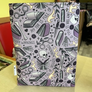 Puede incluir: Una funda de libro morada con un patrón repetido de libros, calaveras, dagas, esposas, huesos y llamas. El texto "My TBR is full of trigger warnings" se repite en todo el patrón.