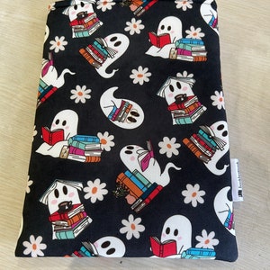Puede incluir: Funda para libro negra con un fantasma blanco leyendo un libro sobre una pila de libros y margaritas blancas. El texto "bookishmark" está en una etiqueta blanca.